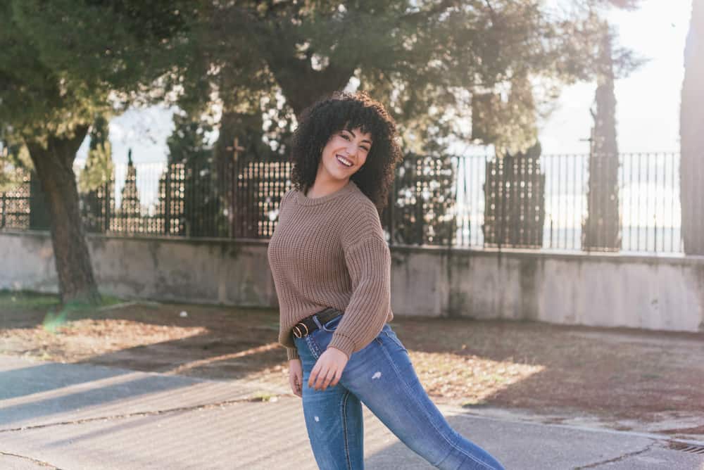 Cute white girl with type 3b curls dyed with old hair dye dancing in the park wearing a beige sweater and torn blue jeans.