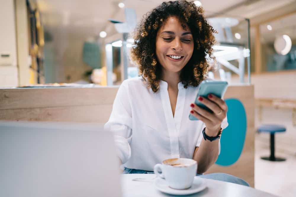 Female sitting in a restaurant having coffee while talking to her husband on the phone, wearing a white shirt and stylish jeans.