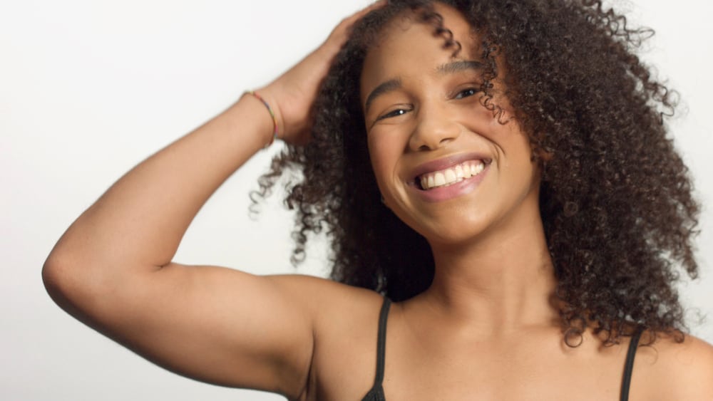 Black woman with curly hair strands rubbing her hands through her hair wearing a multi-colored bracelet, pink lipstick, and a black tank top.