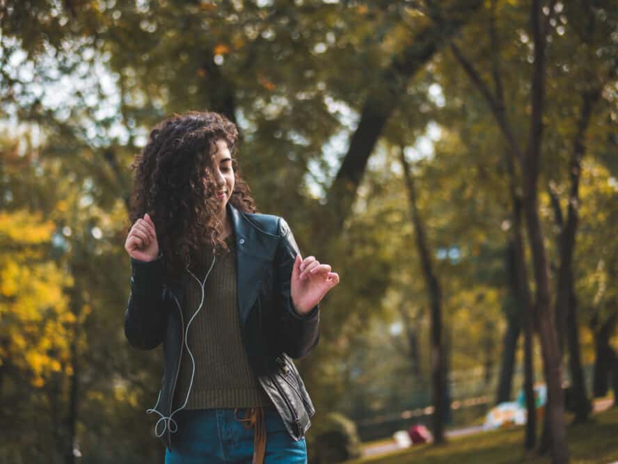 Mixed-raced female dancing outside while listening to music on earphones while wearing a black jacket, green sweater, and blue jeans.