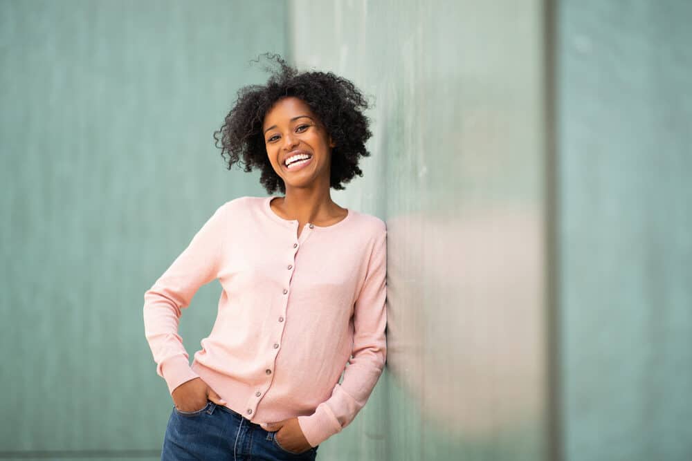 Black woman with 4d hair strands wearing a pink shirt that buttons down the middle and blue jeans. 
