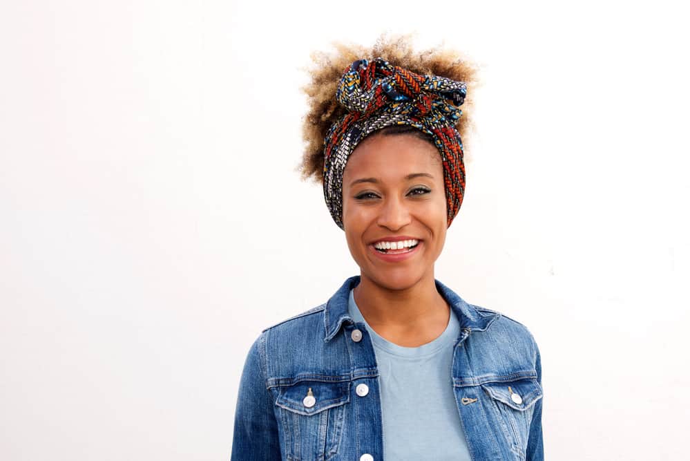 Black lady with bleached hair wearing a headscarf, a blue-jeaned jacket, and a blue t-shirt.