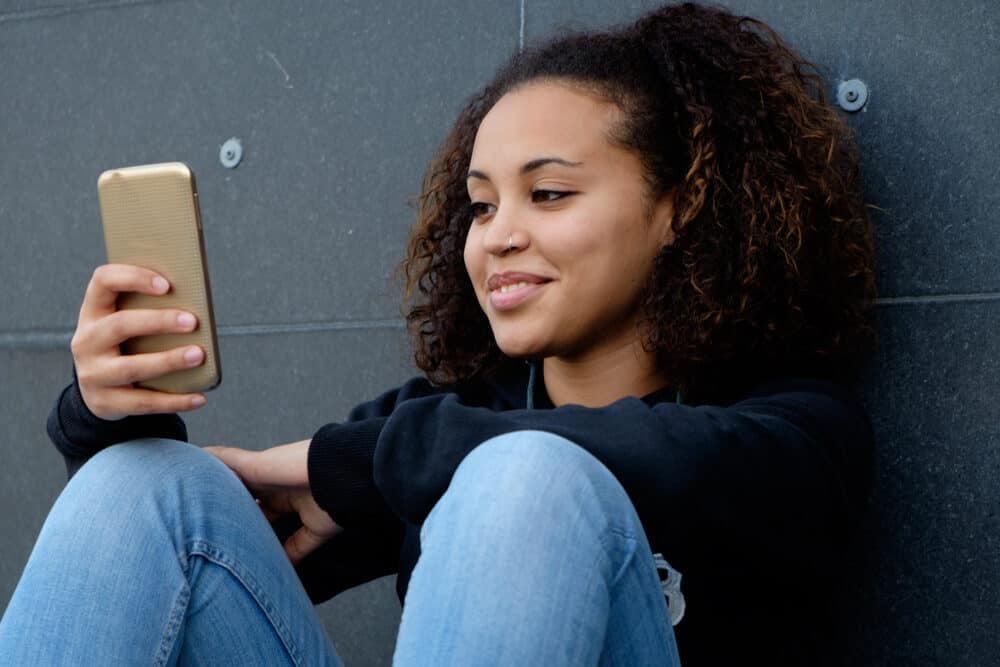 Pretty African American female with curly hair and a nose ring sitting on the ground using her mobile phone. 