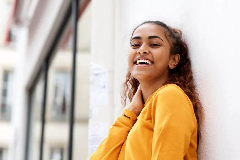 Should You Wash Your Hair Before a Haircut or Before You Color It? 6 Female of Indian descent wearing a yellowish orange shirt standing beside a white wall