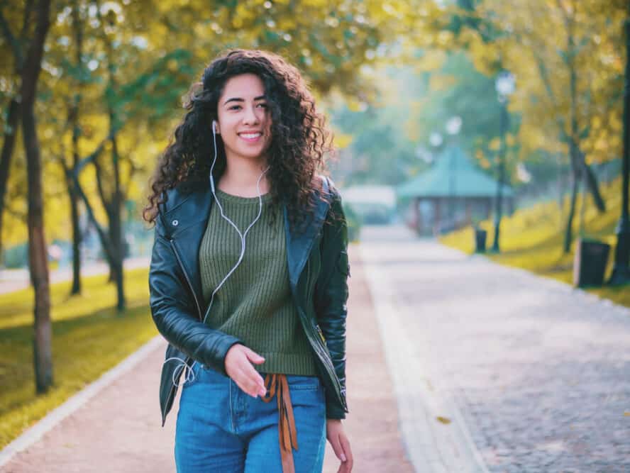 Smiling mixed race female with wavy hair wearing a black leather jacket during the fall season while listening to songs on Apple earphones.