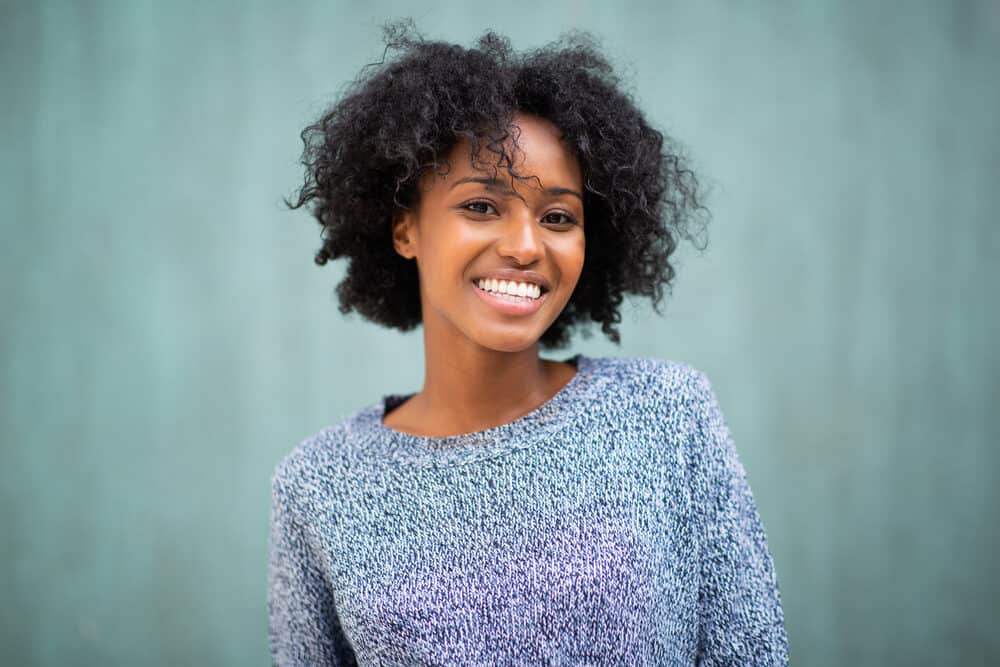 Black women with 4a hair strands smiling standing against a green wall wearing a blue and white sweater.