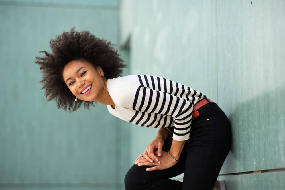 Young black women with 4b hair strands after using a tea tree scalp treatment standing against a wall wearing red lipstick, earrings, a blue and white sweater, red belt, and black jeans.