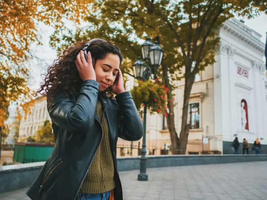 Latin women with naturally curly hair listening to music on Bose headphones wearing a green shirt, blue jeans, and a black leather jacket.
