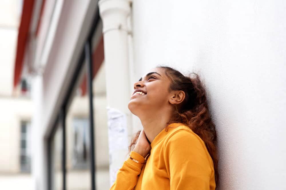 Indian female wearing an orange sweater and a gold braclet leaing against a concrete wall