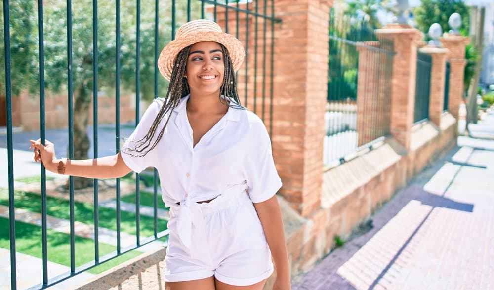 A black woman with braids is standing outside on a sunny day in the summer wearing a white jumper leaning against a black cast iron fence.