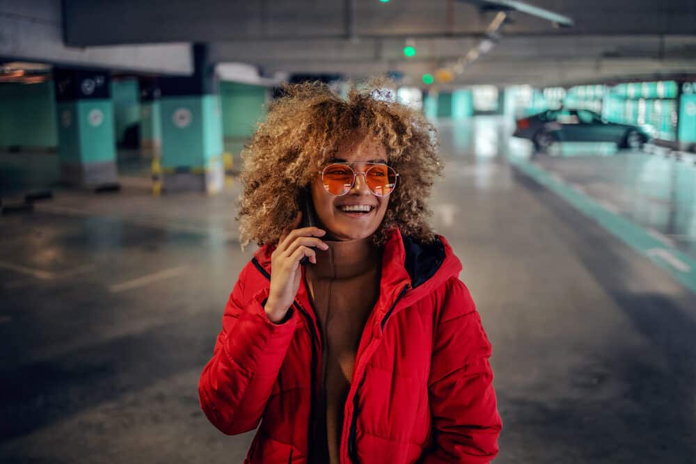 A young woman with permanent hair color in her curly hair, carrying a phone to her ear as she stands in the underground garage.