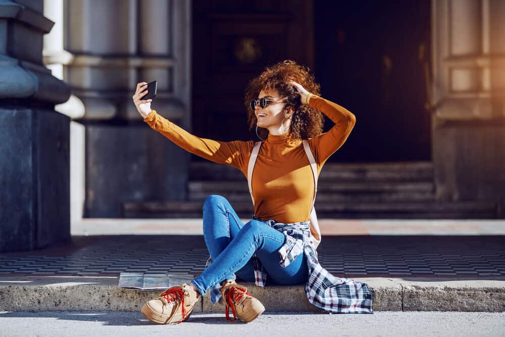 A young woman with curly hair takes a selfie while sitting on the ground outside.