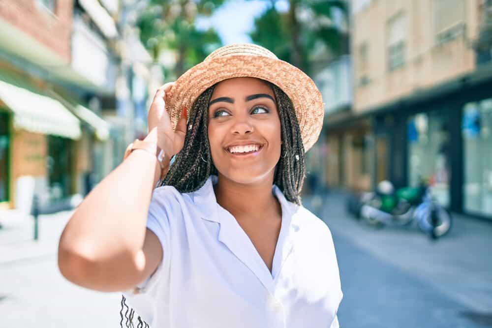 A black woman with braids standing outside on a sunny day in the summer wearing a white jumper and brown straw hat.