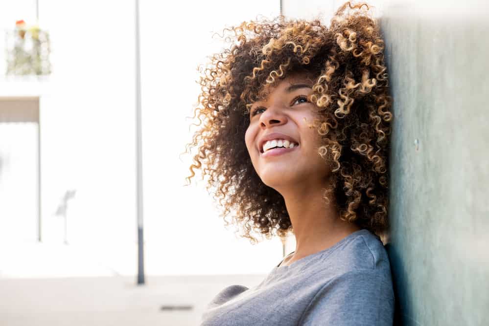 Black girl with ombre curly hair follicles leaning against a stone wall, looking towards the sky while smiling.