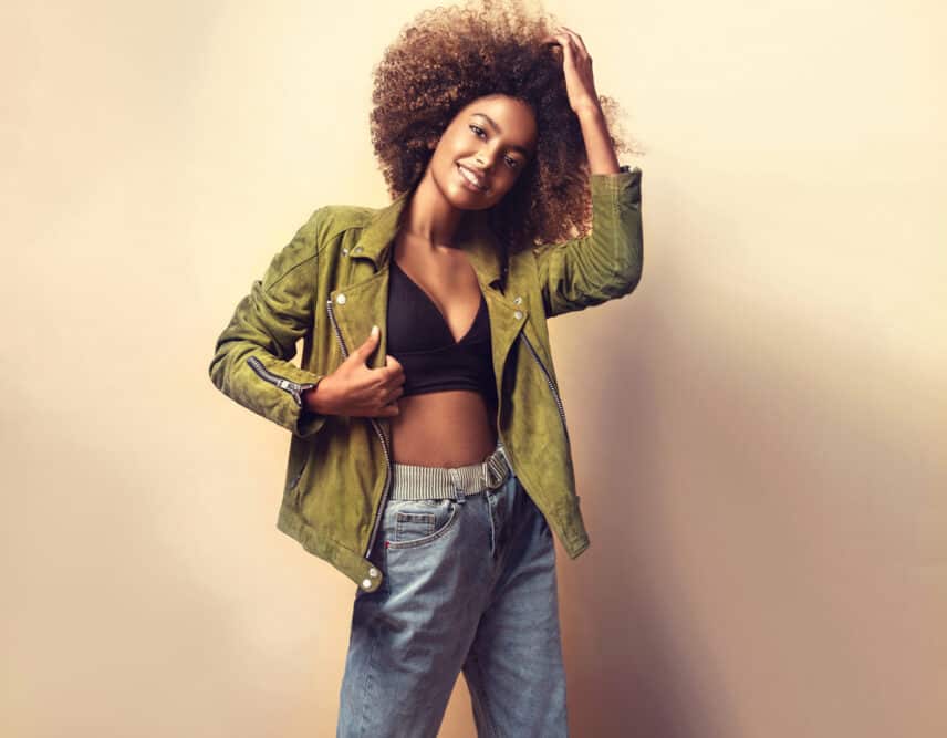 African American women wearing a green jacket, black shirt, blue jeans, and naturally curly brown hair strands.