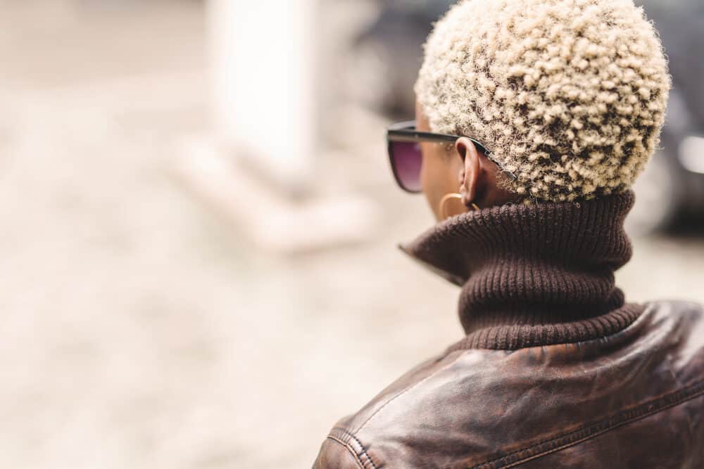 Black woman with a blonde curly afro hairstyle wearing a brown leather coat, hoop earrings, and glasses