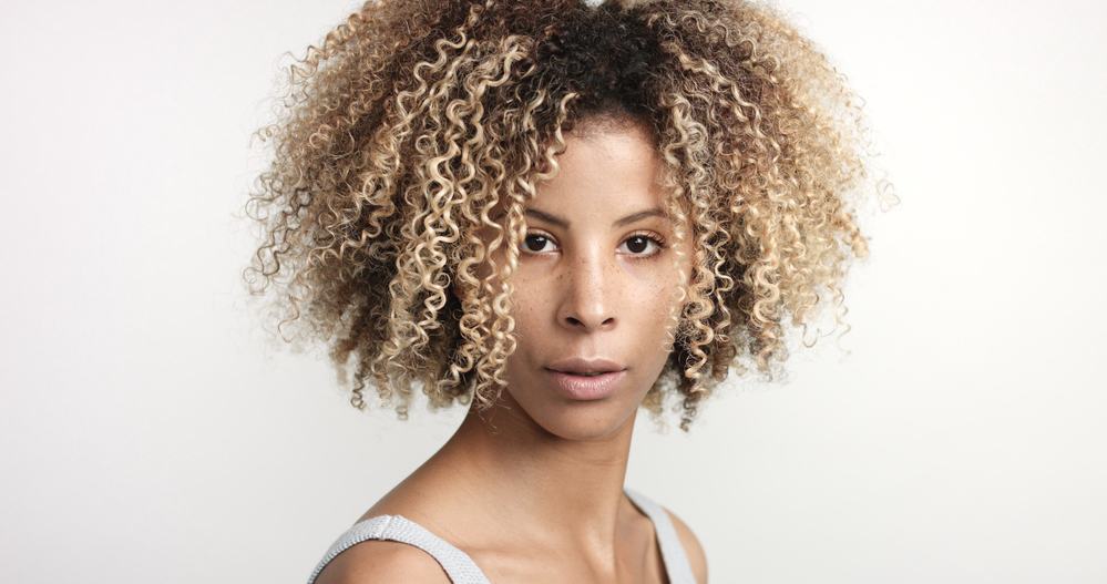 A black woman who has curly blonde hair that covers most of her face is looking up from a sink