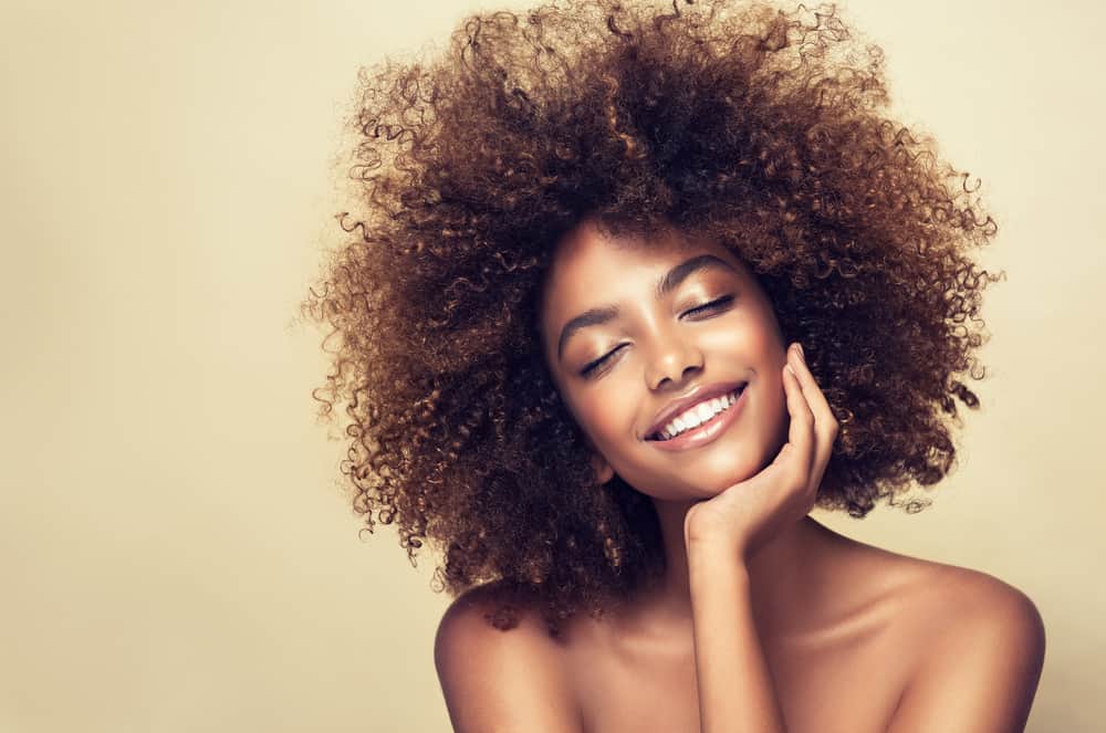 A girl with brown curly hair, pink lipstick and arched eyebrows holding her chin and smiling.