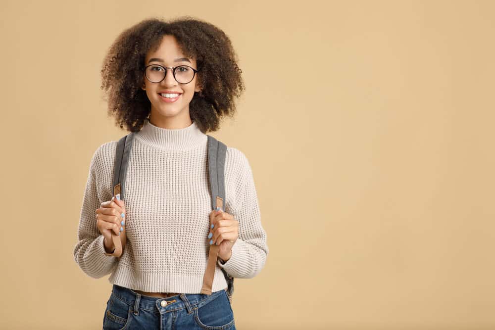 Pretty African American woman with 4a hair strands wearing black glasses, a brown sweater, blue jeans, and a backpack.