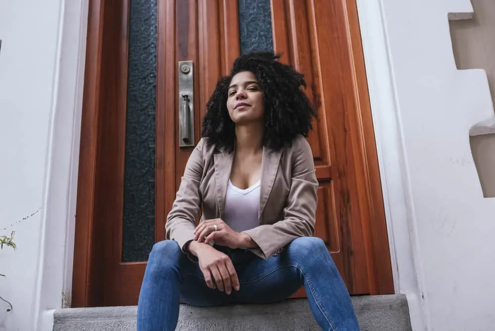 Light skinned female sitting outside wearing blue jeans, a white shirt, brown jacket, and curly natural hair