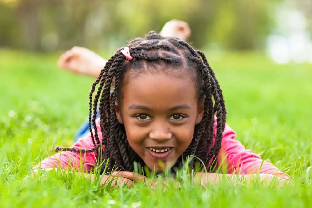 Little diva with brown skin wearing a pink shirt and jeans lying on the ground with braided hair in its natural curly state.