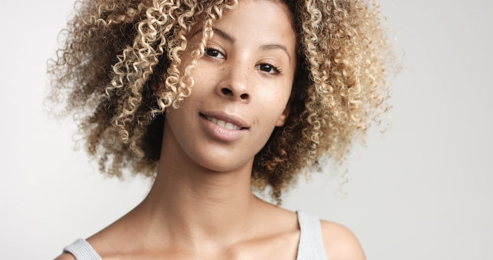 Black women with naturally curly blonde hair styled with natural products wearing a gray tank top and pink lipstick