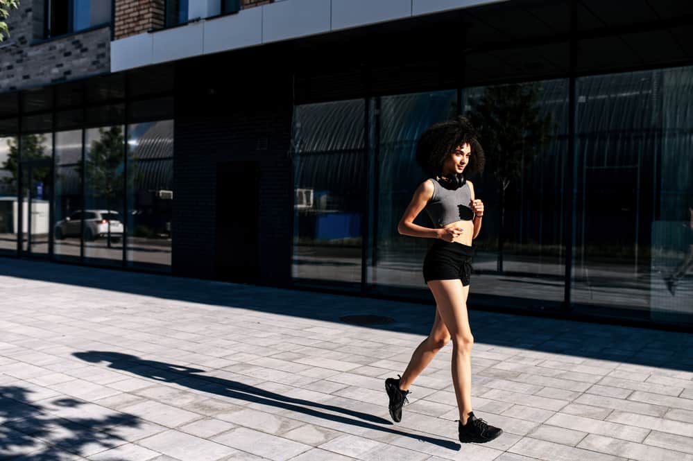 Fit woman with black hair jogs down the street in her workout clothes and headphone.