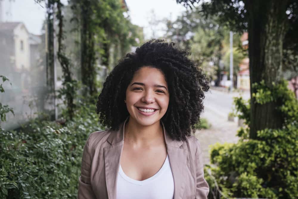 Cute Mexican lady with amazing 3c natural curls and black eyes wearing a white shirt, brown jacket, and pink lipstick