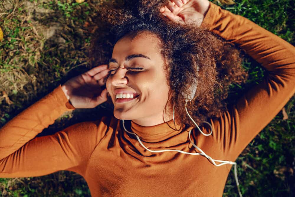 A beautiful, mixed-race woman with curly hair cuddles up to the sound of her headphones and enjoys a day in the park.