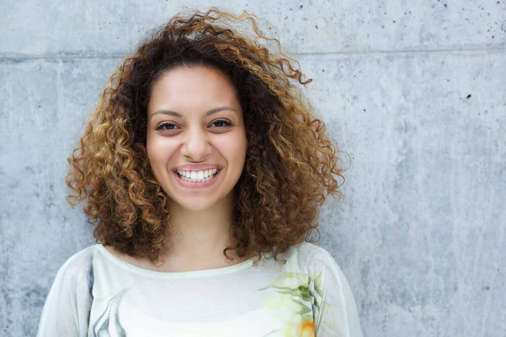 Black girl with natural curls leaning up against a gray wall wearing a green dress shirt with a flower on the left shoulder