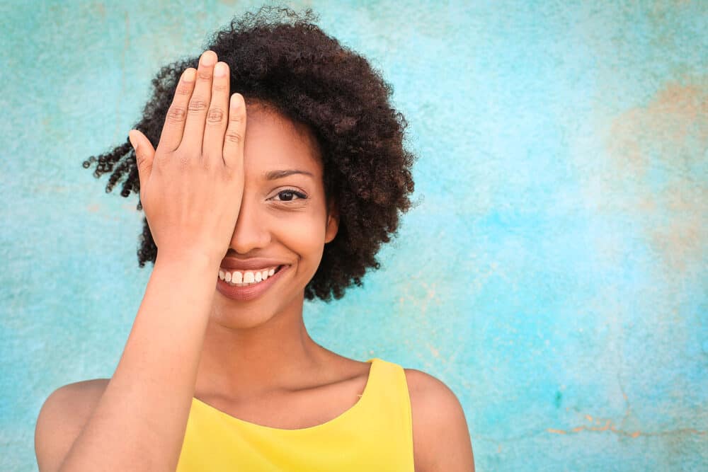 African American female with curly flat hair after using Brazil nut shampoo wearing a yellow shirt while covering her right eye.