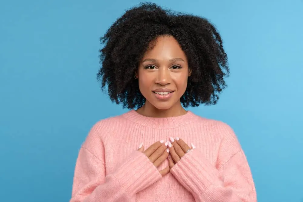 Black girl with curly hair wearing a pink sweater, red lipstick, and pearl-colored fingernail polish.