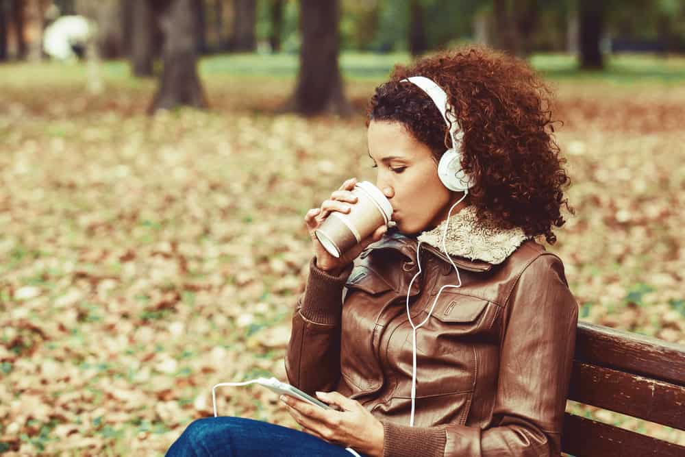 ženy s 3C přírodními vlasy sedí v parku na sobě hnědý kožený kabát a pití kávy.