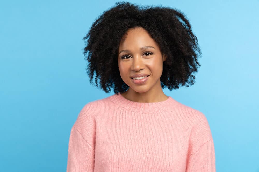 Beautiful woman with curls treated with almond oils smiling wearing a casual pink sweater. 