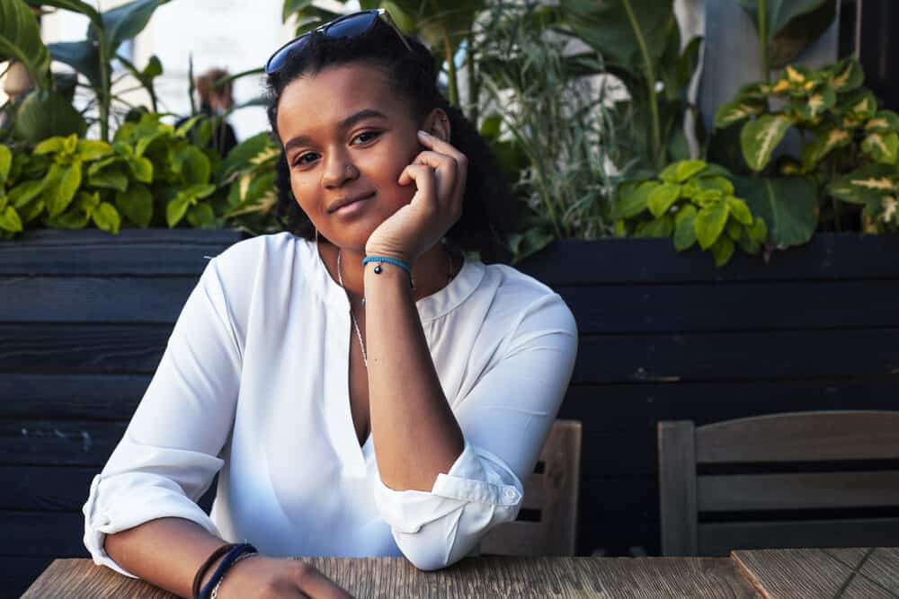 A black lady sitting outside at a restaurant wearing a white shirt, nose ring, with curly hair longer than her shoulders.