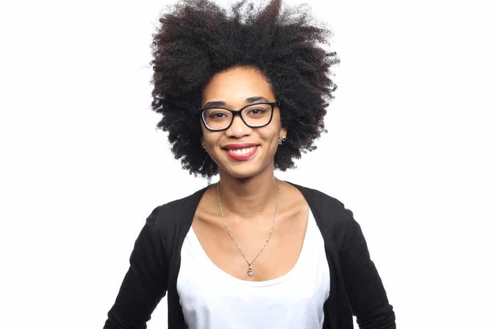An African American female with 4c kinky, coily hair stands wearing black pearl earrings and a white t-shirt.