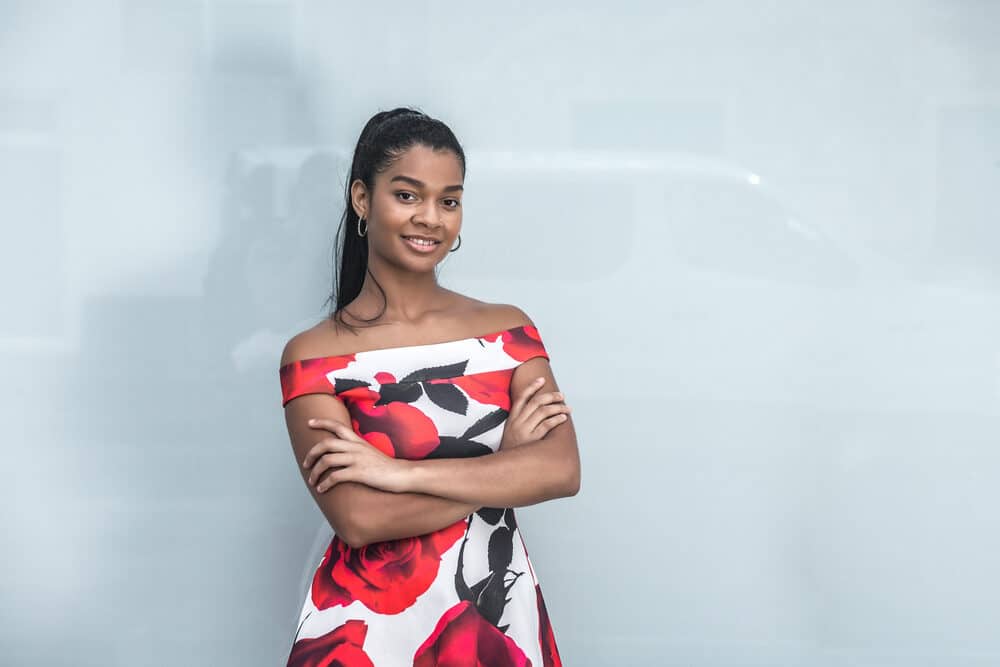 Black lady ready for the cover of Beauty News posing for a photo indoors with her arms folded wearing a colorful dress and a long ponytail on straightened hair.