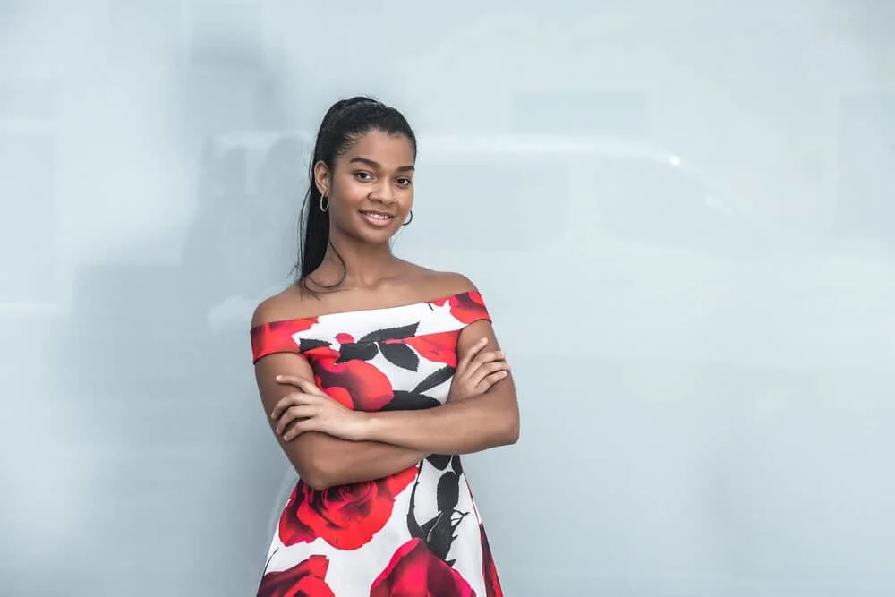How To Do a Sleek Ponytail on Natural Hair (or Weave) Without Flyaways 5 Black lady ready for the cover of Beauty News posing for a photo indoors with her arms folded wearing a colorful dress and a long ponytail on straightened hair.