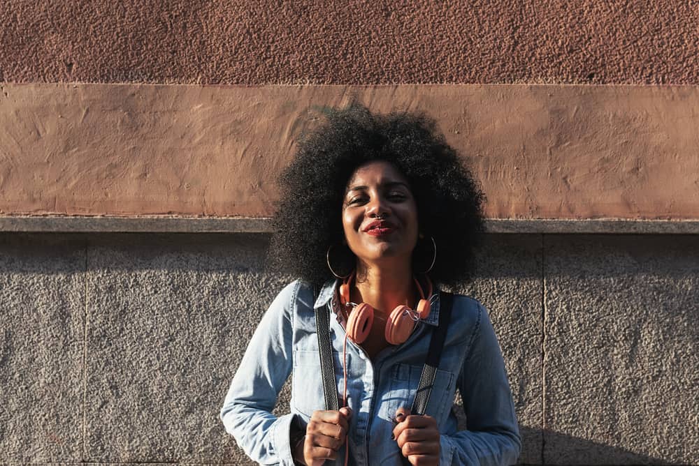 Black girl standing outside laughing while wearing a blue jean shirt and peach headphones.