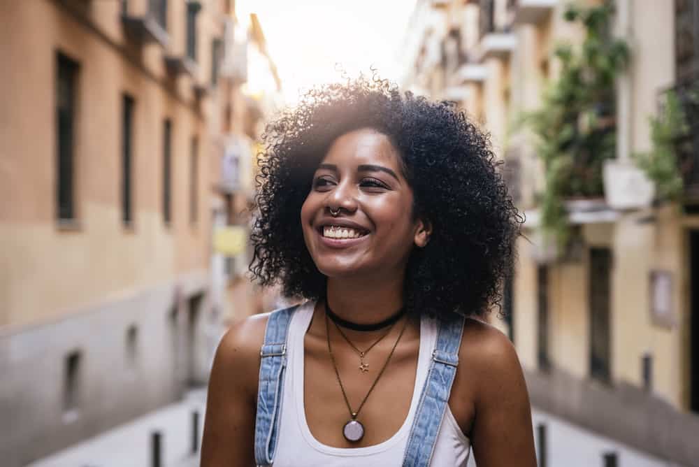 African American female with natural hair wearing a white t-shirt, blue jean overalls, three necklaces, and a nose ring with a big smile.