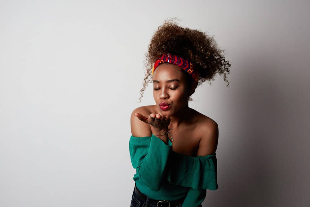 Young black girl with coiled hair wearing an African scarf, red lipstick, and green shirt blowing a kiss to the camera.