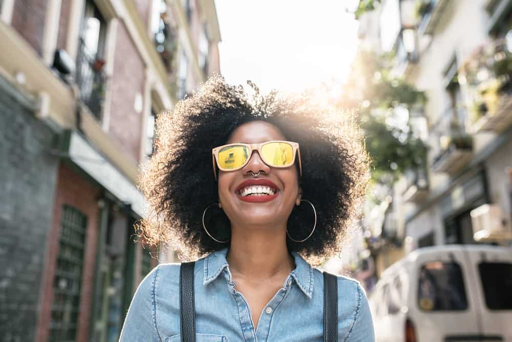 African American female with a natural hairstyle wearing a blue jean shirt, peach sunglasses, and red lipstick.