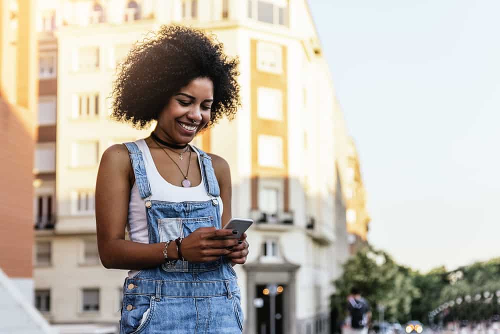 African American woman with curly hair wearing a white t-shirt, blue jeans, and three necklaces using an Apple mobile phone.