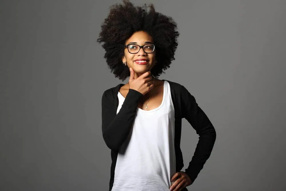 What is Coily Hair and How Do I Care for It? 4 A black woman with type 4c hair holding her chin wearing a white T-shirt, black glasses, earrings, and red lipstick.