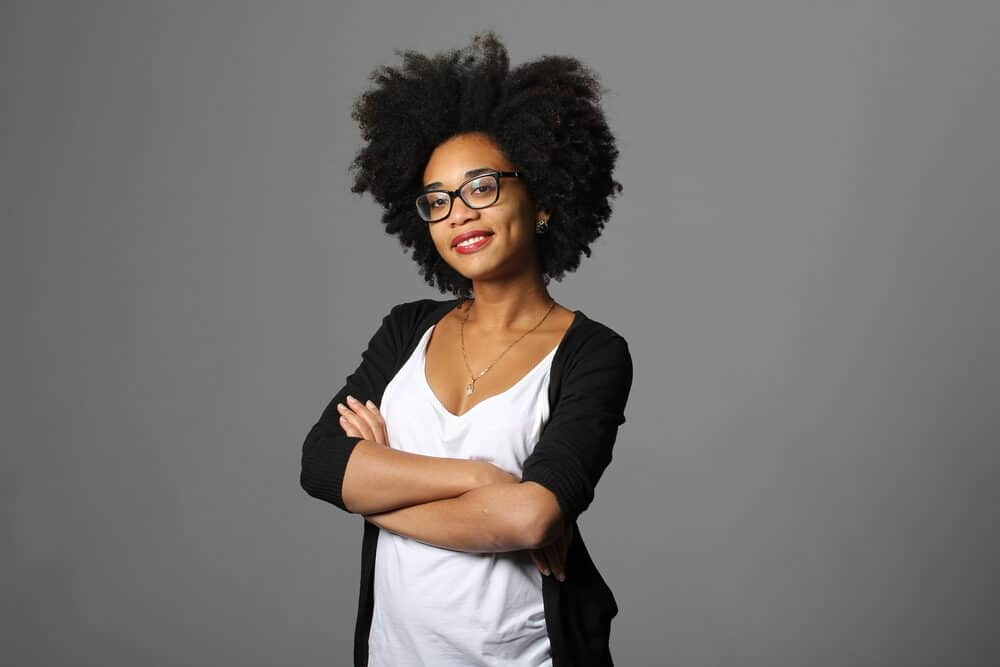 African American woman with her arms crossed wearing a white t-shirt, black glasses, earrings, kinky coily hair strands, and red lipstick.