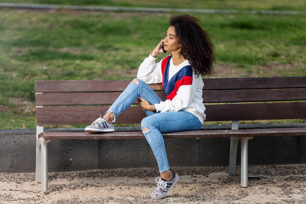 African lady with virgin hair wearing curly weave while sitting on a park bench talking on a mobile phone.