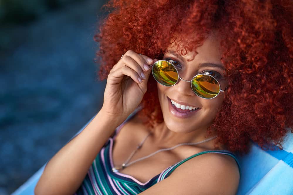 Women with red curly hair extensions treated with purple shampoo wearing sunglasses by the pool side.