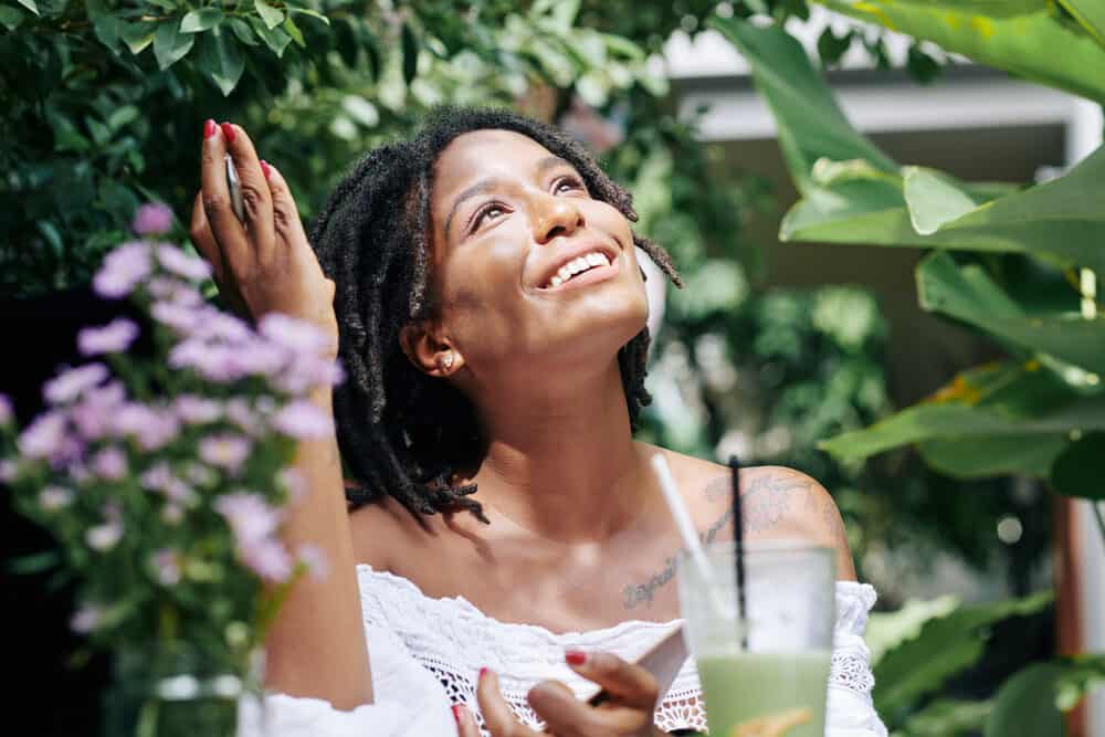 A cute woman with 4c dreads looking at the sky and smiling while outside with plants in the background.