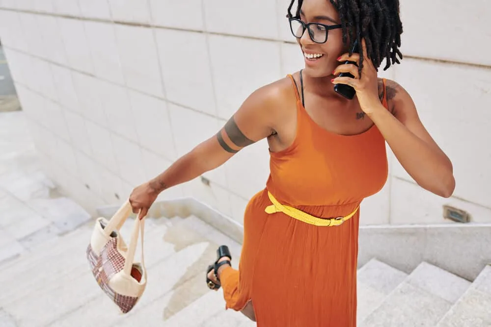 A young African woman in a bright orange dress talks on the phone about using natural scalp oil for healthier hair growth.