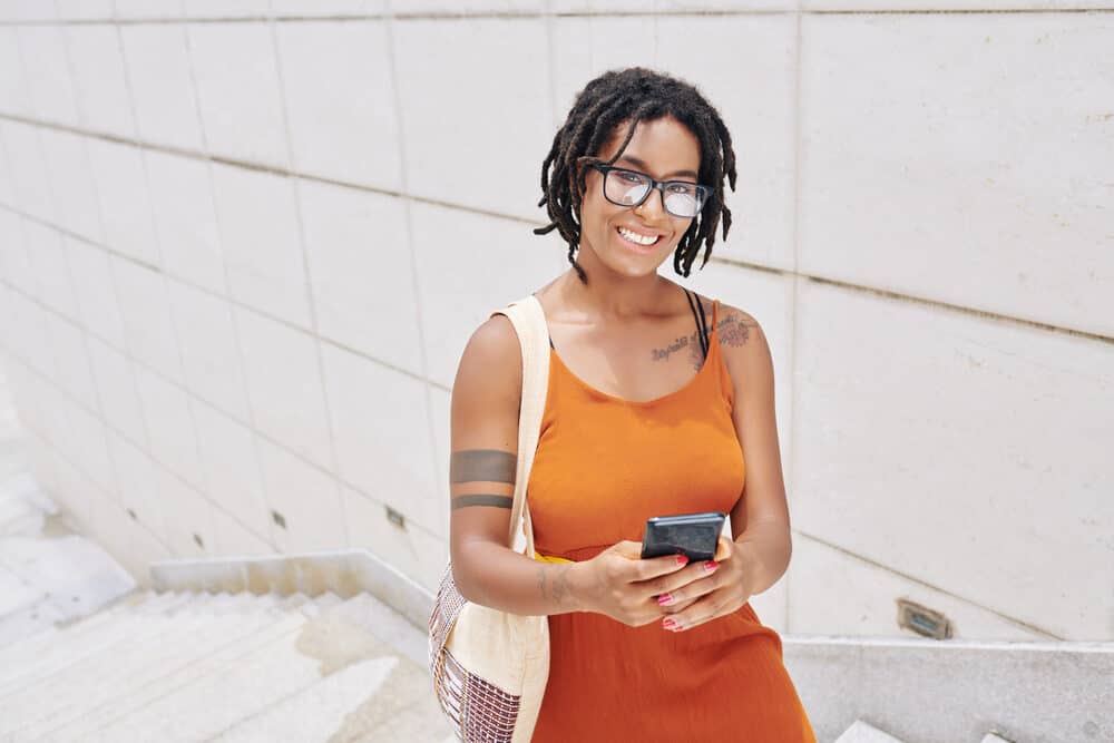 A young African woman with glasses and a sophisticated dreadlocks hairstyle smiles at the camera while she uses her mobile phone outdoors.